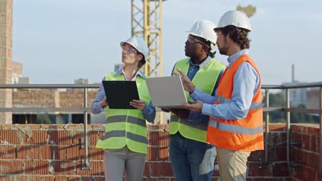 Three-young-mixed-races-builders-and-architects,-two-men-and-woman-standing-together-at-the-constructing-site-with-laptop-computer-and-tablet-device-and-discussing-work-process.